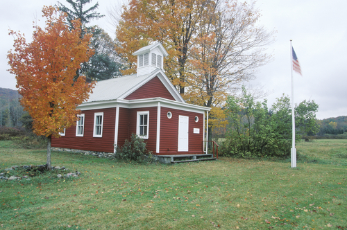 Red one-room schoolhouse with a bell cupola and American flag among autumn trees