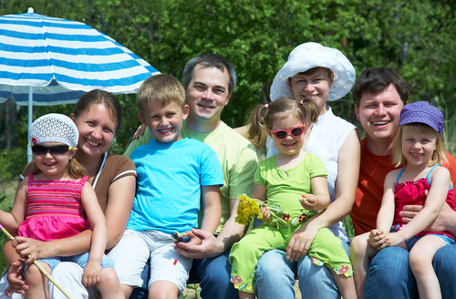 Extended family with young children smiling outdoors under a striped umbrella