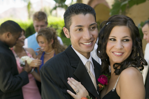 Teenage couple dressed for prom smiling outdoors, friends behind them