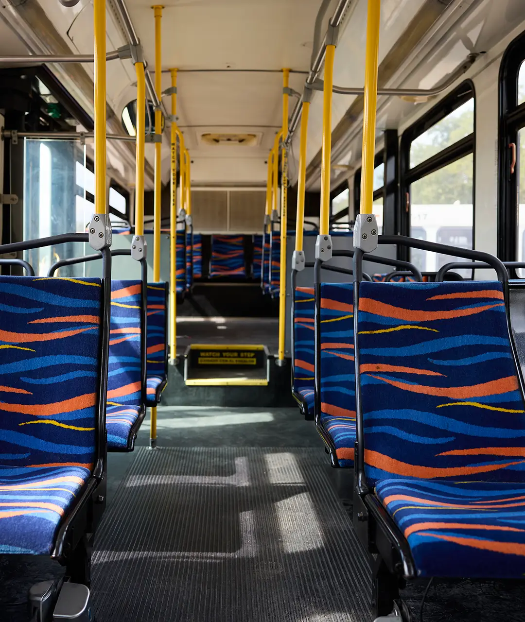 Empty interior of a First Class Transportation transit bus looking down the center aisle, with blue and orange wave-patterned seats and yellow grab poles