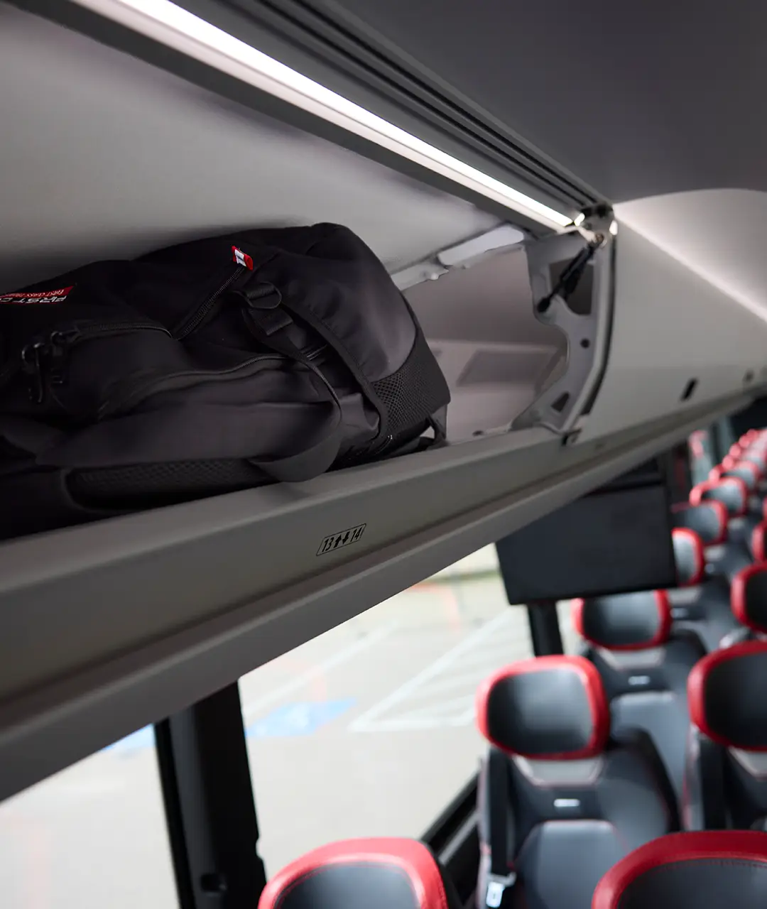 Black backpack stowed in the overhead luggage rack above red-trimmed seats on a First Class Transportation motorcoach
