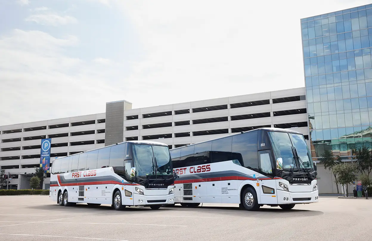 Two white First Class Transportation full-size motorcoaches parked side by side outside a parking garage