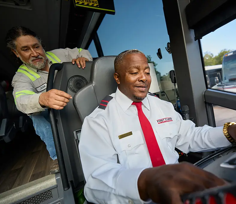 First Class Transportation driver seated at the wheel of a motorcoach with a technician standing behind