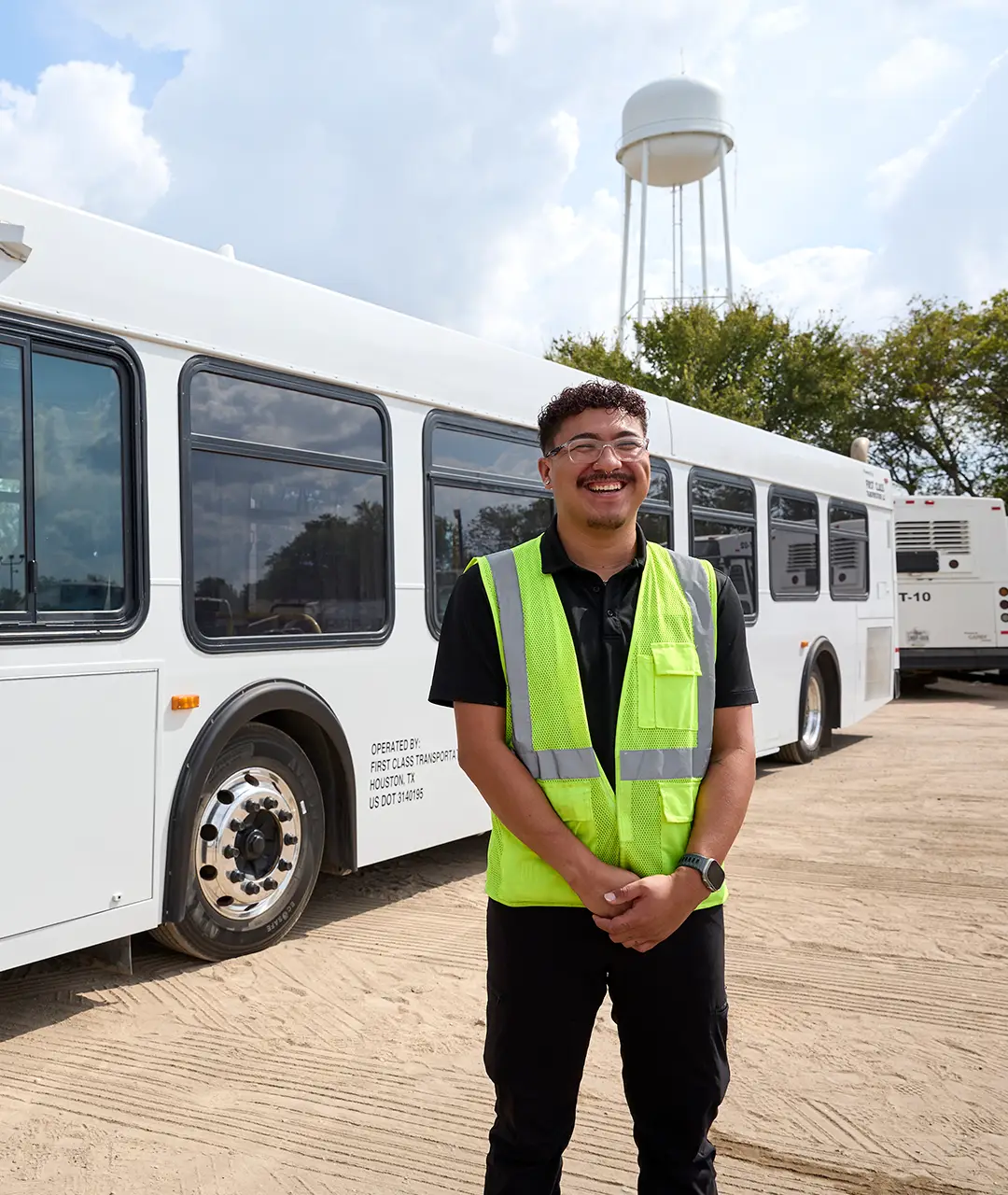 First Class Transportation employee in a hi-vis safety vest standing in front of a white transit bus in the company lot