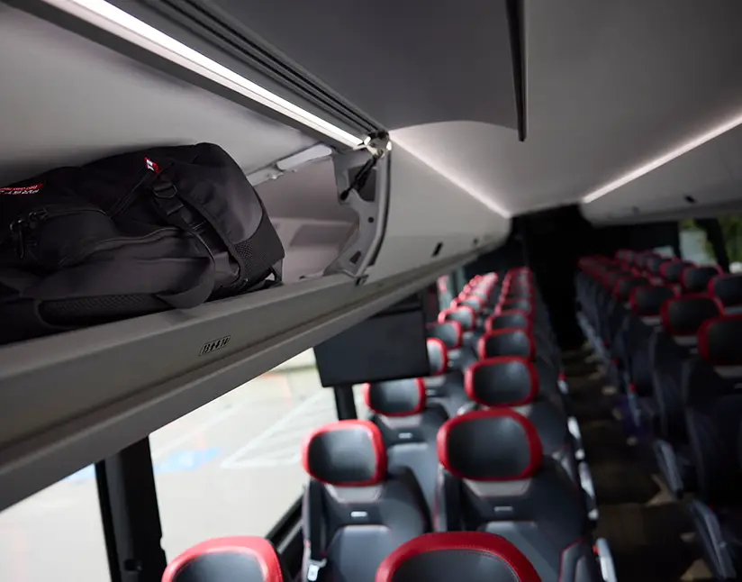 Open overhead luggage compartment holding a bag above First Class Transportation motorcoach seats