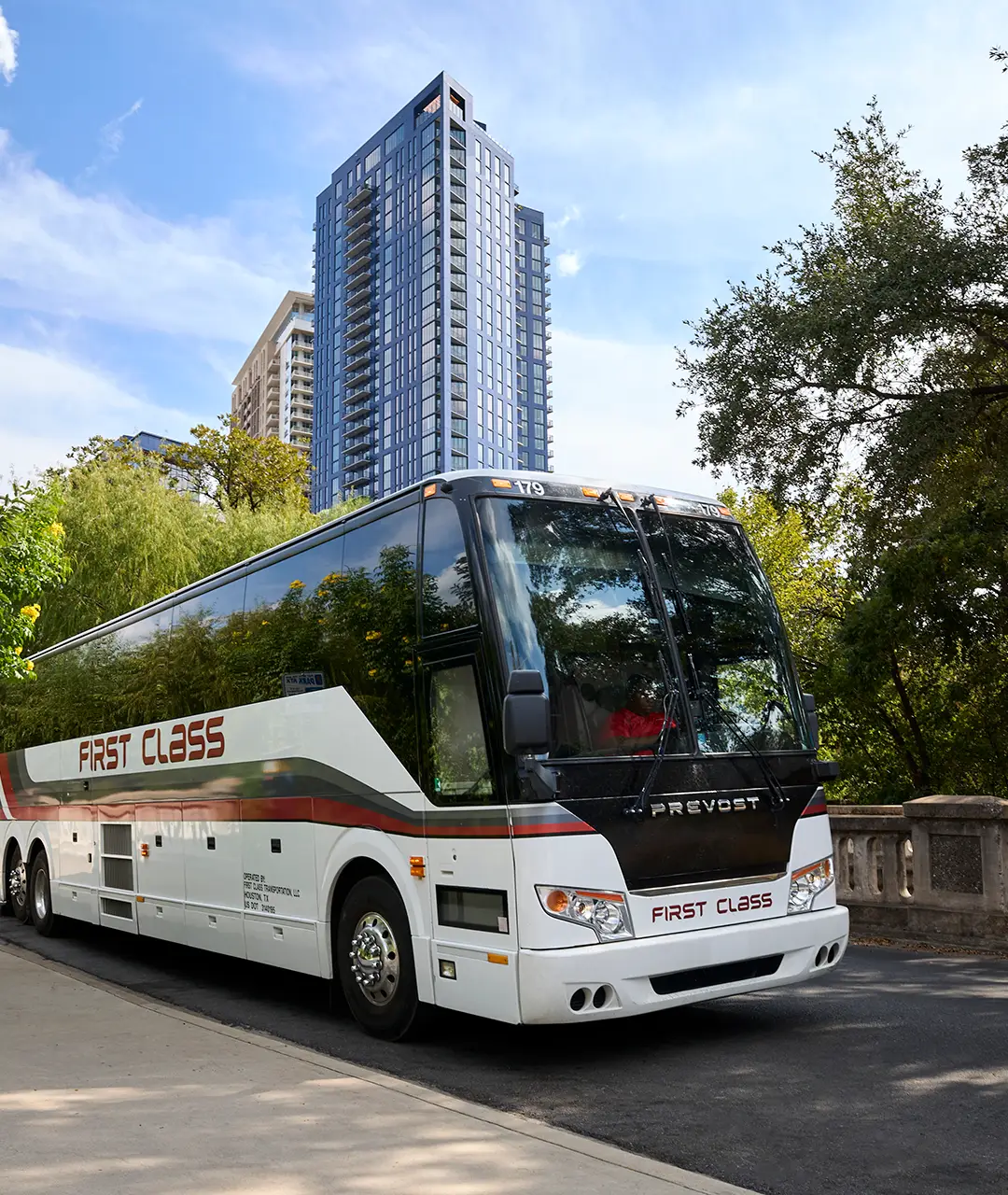 White First Class Transportation Prevost motorcoach with red First Class lettering driving past a city high-rise