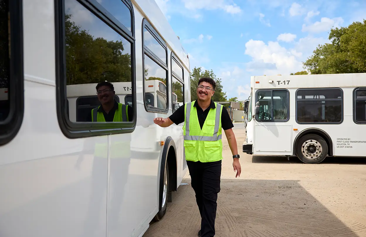 First Class Transportation team member in a safety vest walking between white transit buses at the depot