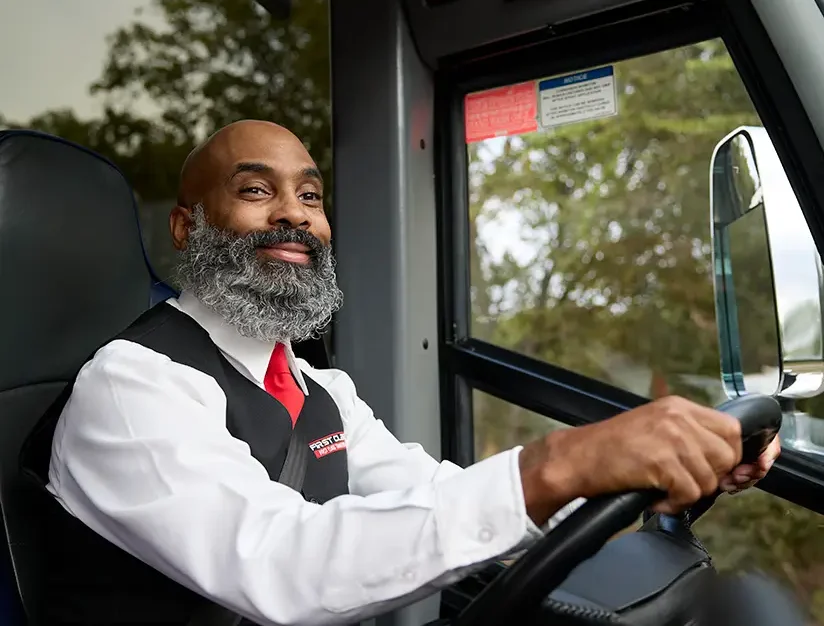 Smiling First Class Transportation bus driver in a black vest and red tie seated at the steering wheel